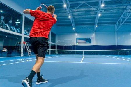 Téléchargez les photos : Jeune homme sur un court de tennis intérieur frappant la balle avec une raquette servant pendant le sport professionnel instructeur de tennis de match - en image libre de droit