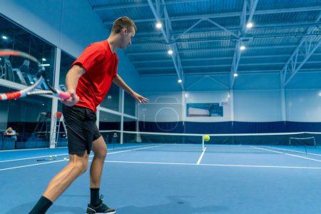 Foto de Joven en una cancha de tenis cubierta golpeando la pelota con una raqueta que sirve durante el juego instructor de tenis deporte profesional - Imagen libre de derechos