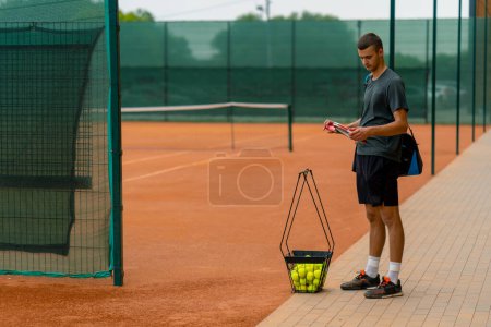 Foto de Entrenador joven jugador profesional de pie en la cancha de tenis al aire libre antes de comenzar el entrenamiento con raqueta y balones de tenis cesta - Imagen libre de derechos