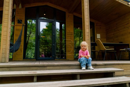 Téléchargez les photos : Petite fille mignonne et souriante dans des vêtements élégants sur la terrasse d'une maison en bois dans les bois - en image libre de droit