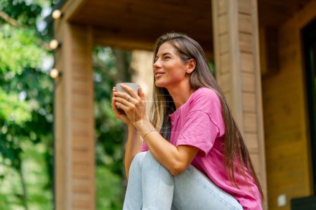 Téléchargez les photos : Une fille réfléchie s'assoit avec une tasse de thé sur la terrasse de son chalet en bois dans les bois le matin - en image libre de droit