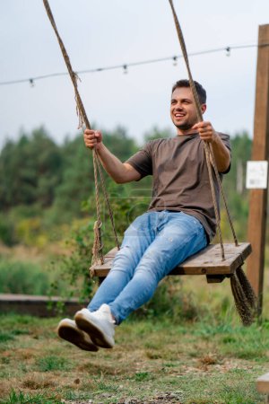 Photo for A young handsome man smiles and rides a large wooden swing in a field in nature overlooking a lake - Royalty Free Image