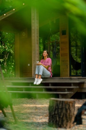 Foto de Una niña pensativa se sienta con una taza de té en la terraza de su casa de madera en el bosque por la mañana - Imagen libre de derechos