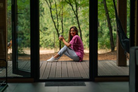 Téléchargez les photos : Une jeune femme souriante s'assoit latéralement sur une terrasse surplombant la forêt et profite du silence - en image libre de droit