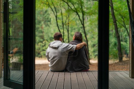 Foto de Hombre y mujer enamorados sentados de espaldas a la cámara en una terraza con vistas al bosque con cuadros abrazándose y hablando - Imagen libre de derechos