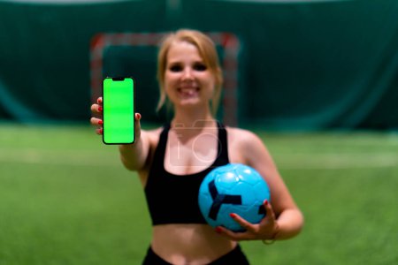 Photo for Smiling young soccer girl showing phone with green screen and holding ball with hand soccer field - Royalty Free Image