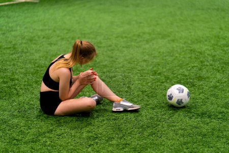 Téléchargez les photos : Jeune fille footballeuse blessé à la jambe lors d'un match d'entraînement assis sur l'herbe vérifier son genou en attente d'aide - en image libre de droit