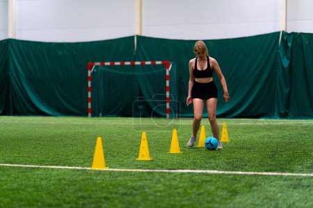 Foto de Niña está entrenando en el campo de fútbol conduciendo la pelota entre las fichas o conos preparándose para el partido - Imagen libre de derechos