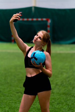 Foto de Sonriente joven blogger chica de fútbol tomando fotos en el teléfono tomando selfie con campo de fútbol de pelota - Imagen libre de derechos