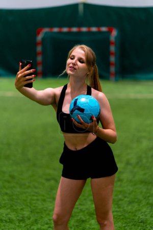 Foto de Sonriente joven blogger chica de fútbol tomando fotos en el teléfono tomando selfie con campo de fútbol de pelota - Imagen libre de derechos