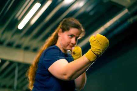 Photo for Portrait focused Boxer girl in yellow boxing bandages standing in a boxing stance on a dark background in gym before a fight - Royalty Free Image