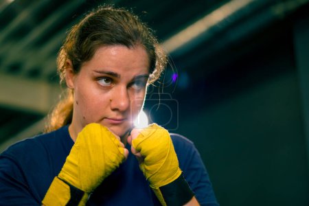 Photo for Portrait focused Boxer girl in yellow boxing bandages standing in a boxing stance on a dark background in gym before a fight - Royalty Free Image