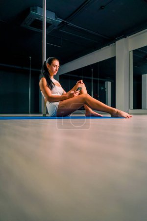 Photo for Portrait of a young beautiful sexy girl after a dance class on a pole holding phone in her hands texting with someone - Royalty Free Image