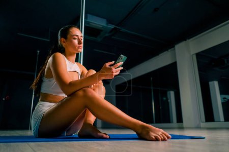 Photo for Portrait of a young beautiful sexy girl after a dance class on a pole holding phone in her hands texting with someone - Royalty Free Image