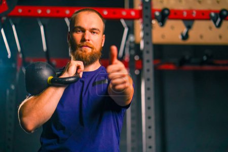 Photo for Portrait of a satisfied athlete in the gym after a workout a man shows a super sign with his hand calls for a sporty lifestyle holding kettlebell - Royalty Free Image