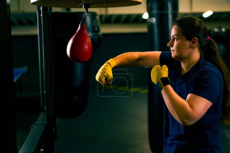 Foto de Una chica boxeadora con guantes resuelve los golpes de fuerza y la coordinación en un pequeño saco de boxeo en el gimnasio entrena diligentemente antes de la pelea - Imagen libre de derechos