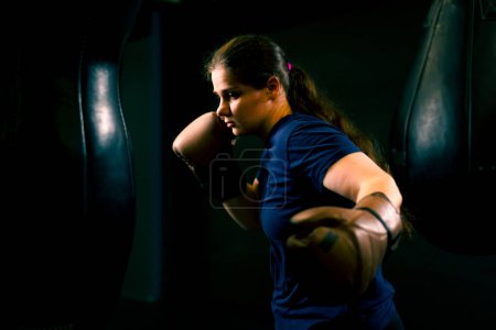 Téléchargez les photos : Un boxeur de fille dans des gants travaille sur la puissance des coups de poing sur un sac de boxe dans les trains de gymnastique dur avant de se battre - en image libre de droit