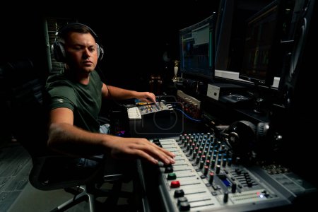 Foto de Un ingeniero de sonido masculino enfocado que usa auriculares se sienta en un escritorio de mezcla grabando una nueva pista - Imagen libre de derechos