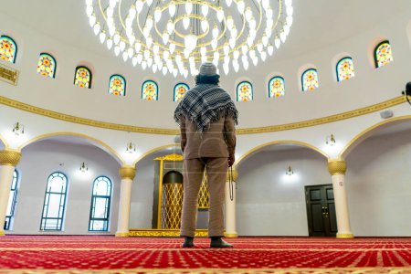 Foto de Disparo por la espalda Un hombre musulmán se para en la alfombra en medio de la sala de oración en la mezquita con la cabeza levantada - Imagen libre de derechos