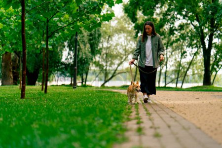 Téléchargez les photos : Jeune fille dans le parc sur une promenade tenant une laisse avec petit chien rouge soins et amour pour les animaux - en image libre de droit