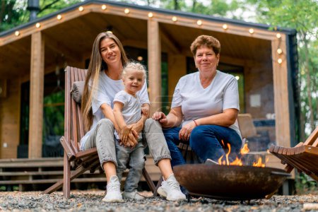 Foto de Generación de mujeres hija madre y abuela sentadas juntas junto al fuego cerca de una casa de madera en la naturaleza - Imagen libre de derechos