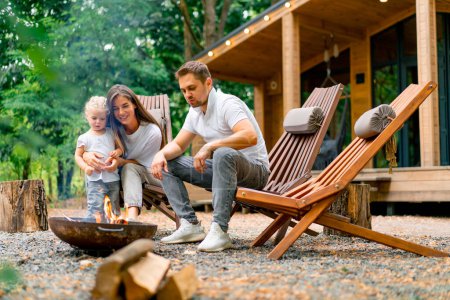Téléchargez les photos : Mignon jeune famille papa maman et sa fille sont assis ensemble par un feu dans la forêt et rôtissant des guimauves près de leur maison de campagne - en image libre de droit
