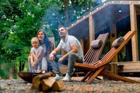 Foto de Lindo padre de familia joven mamá e hija están sentadas juntas junto a un fuego en el bosque y asando malvaviscos cerca de su casa de campo - Imagen libre de derechos