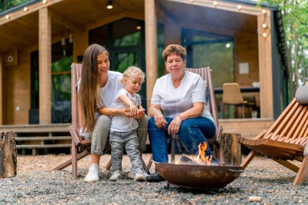 Téléchargez les photos : Génération de femmes fille mère et grand-mère assises au coin du feu près d'une maison en bois dans la nature - en image libre de droit