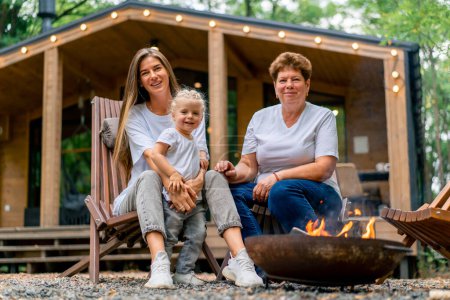 Photo for Generation of women daughter mother and grandmother sitting together by the fire near a wooden house in nature - Royalty Free Image