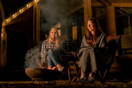 Foto de Novias en mantas se sientan por la noche junto al fuego cerca de una casa de madera del campo en el bosque - Imagen libre de derechos