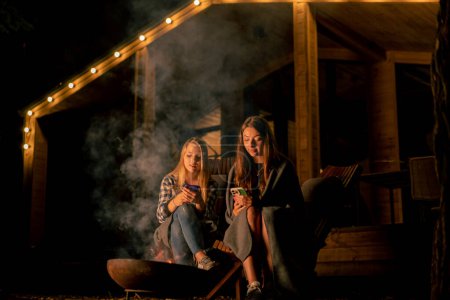 Photo for Girlfriends in blankets sit in the evening by the fire near a country wooden house in the forest - Royalty Free Image