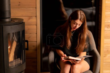 Téléchargez les photos : Une chère fille s'assoit près de la cheminée le soir et lit attentivement un livre dans une maison en bois - en image libre de droit