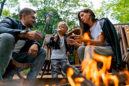 Photo for Cute young family dad mom and daughter are sitting together by a fire in the forest and roasting marshmallows near their country house - Royalty Free Image