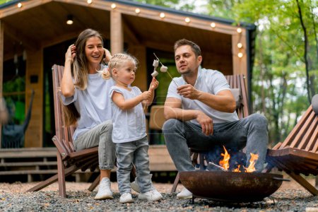Photo for Cute young family dad mom and daughter are sitting together by a fire in the forest and roasting marshmallows near their country house - Royalty Free Image