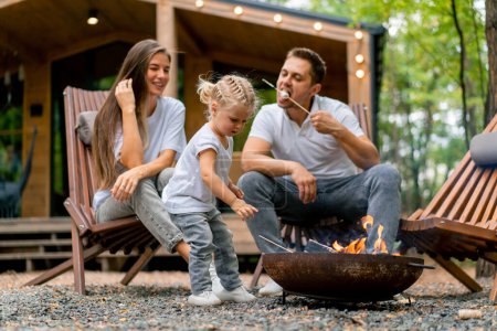 Téléchargez les photos : Mignon jeune famille papa maman et sa fille sont assis ensemble par un feu dans la forêt et rôtissant des guimauves près de leur maison de campagne - en image libre de droit