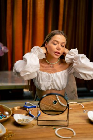 Foto de Retrato de una joven hermosa costurera probándose un collar de perlas alardeando de sí misma en el espejo artesanal producción hobby belleza - Imagen libre de derechos