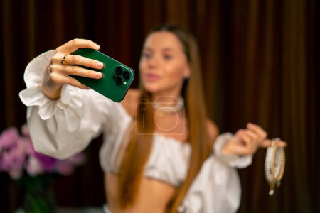 Photo for Portrait beautiful young needlewoman blogger holding a phone in her hands taking a photo of herself with necklace in her hands craft production - Royalty Free Image