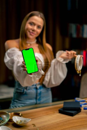Photo for Portrait of a beautiful young needlewoman holding a phone with a green screen and many necklaces craft production hobby work home - Royalty Free Image