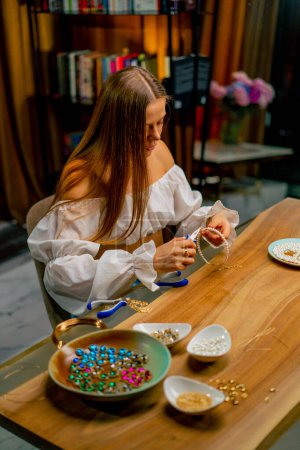 Téléchargez les photos : Une jeune belle fille fait des bijoux avec ses propres mains coupe les restes de fil pour collier avec pinces production artisanale - en image libre de droit