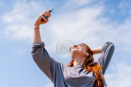 Téléchargez les photos : Une jeune fille rousse en lunettes de soleil avec un téléphone dans ses mains prend une photo d'elle-même sur un appareil photo selfie - en image libre de droit
