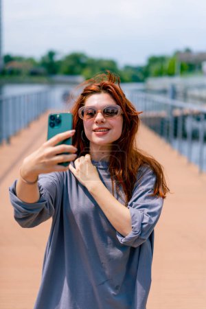 Téléchargez les photos : Une jeune fille rousse en lunettes de soleil avec un téléphone dans ses mains prend une photo d'elle-même sur un appareil photo selfie - en image libre de droit