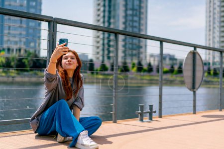 Téléchargez les photos : Jeune fille rousse assis sur une jetée ou un pont avec un téléphone fait blog vidéo dans les réseaux sociaux - en image libre de droit