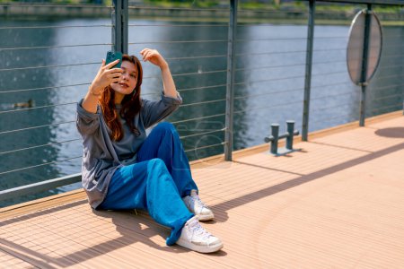 Téléchargez les photos : Jeune fille rousse assis sur une jetée ou un pont avec un téléphone fait blog vidéo dans les réseaux sociaux - en image libre de droit