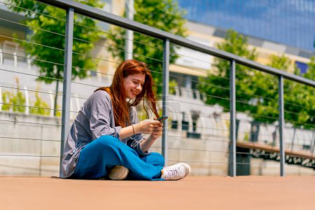 Téléchargez les photos : Une jeune fille rousse est assise sur une jetée ou un pont avec le téléphone et communique avec un ami dans le messager de la communication dans les réseaux sociaux - en image libre de droit