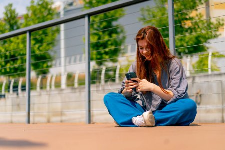 Téléchargez les photos : Une jeune fille rousse est assise sur une jetée ou un pont avec le téléphone et communique avec un ami dans le messager de la communication dans les réseaux sociaux - en image libre de droit