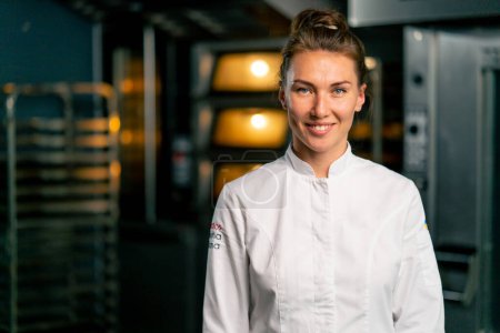 Photo for Portrait of a smiling female chef baker in a professional uniform in a bakery against the background of an oven and fresh baked goods - Royalty Free Image