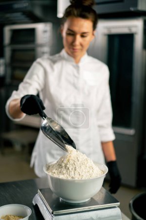 Téléchargez les photos : Gros plan d'une boulangère dans la tunique d'un chef blanc versant de la farine pour préparer et cuire le pain dans une boulangerie - en image libre de droit