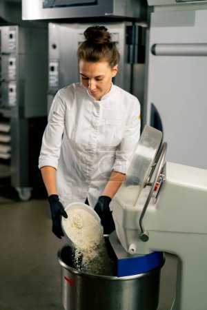 Téléchargez les photos : Une boulangère dans la tunique d'un chef blanc pétrit la pâte pour préparer et cuire le pain dans la boulangerie - en image libre de droit