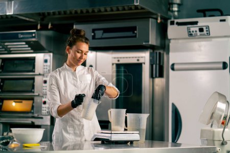 Téléchargez les photos : Femme chef versant de l'eau dans un verre à mesurer debout sur une balance de cuisine dans une cuisine professionnelle - en image libre de droit