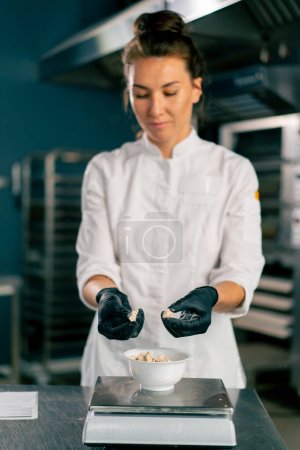 Téléchargez les photos : Une boulangère dans la tunique d'un chef blanc pétrit la pâte pour préparer et cuire le pain dans la boulangerie - en image libre de droit